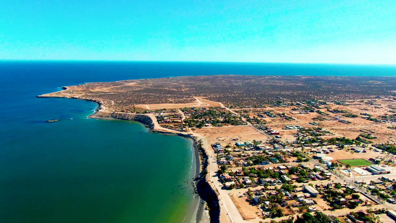 High drone view of a curved bay, shore road, houses, and dry terrain under clear sky.