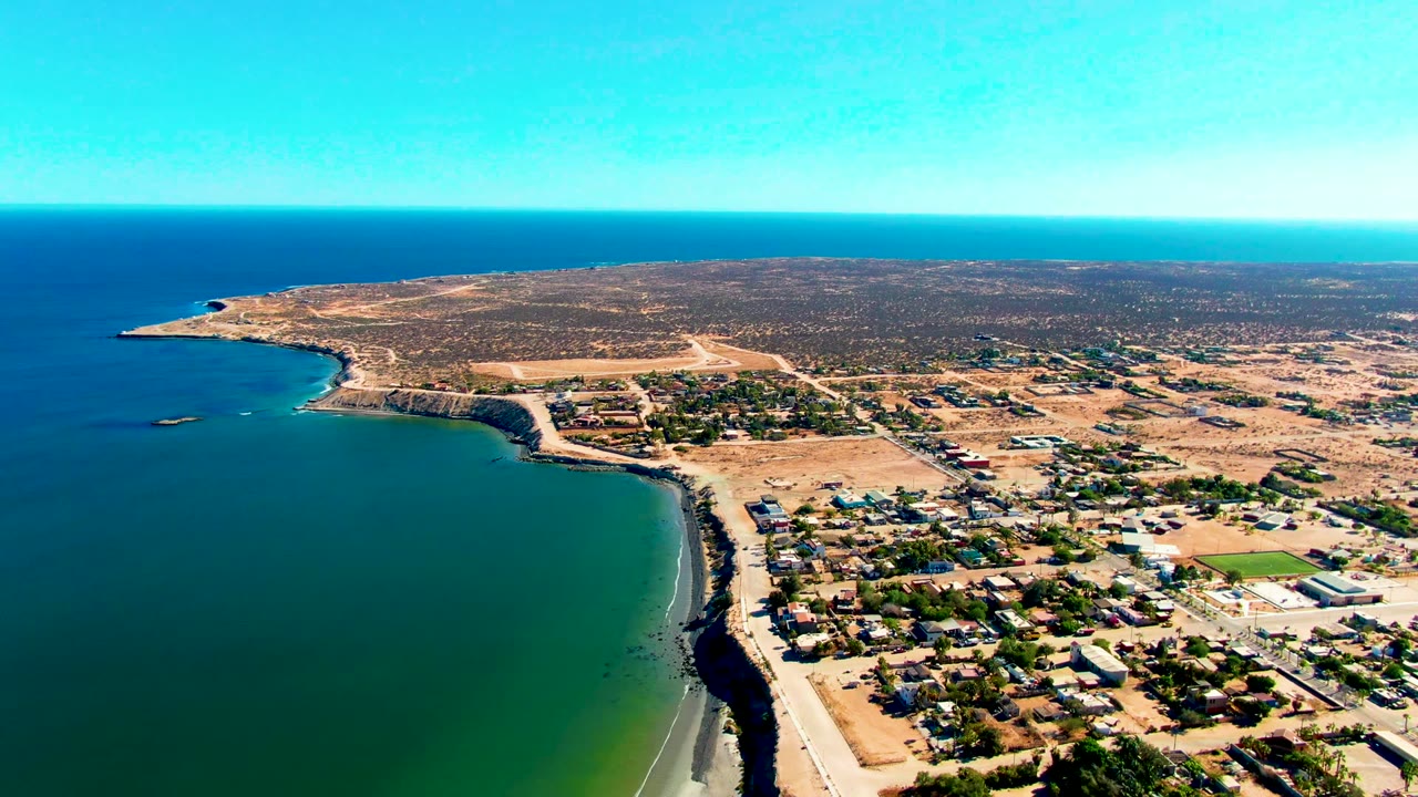 Third aerial frame over San Juanico, Baja California Sur, with turquoise shallows close to shore, town blocks on dry ground, open inland landscape, and no visible people.