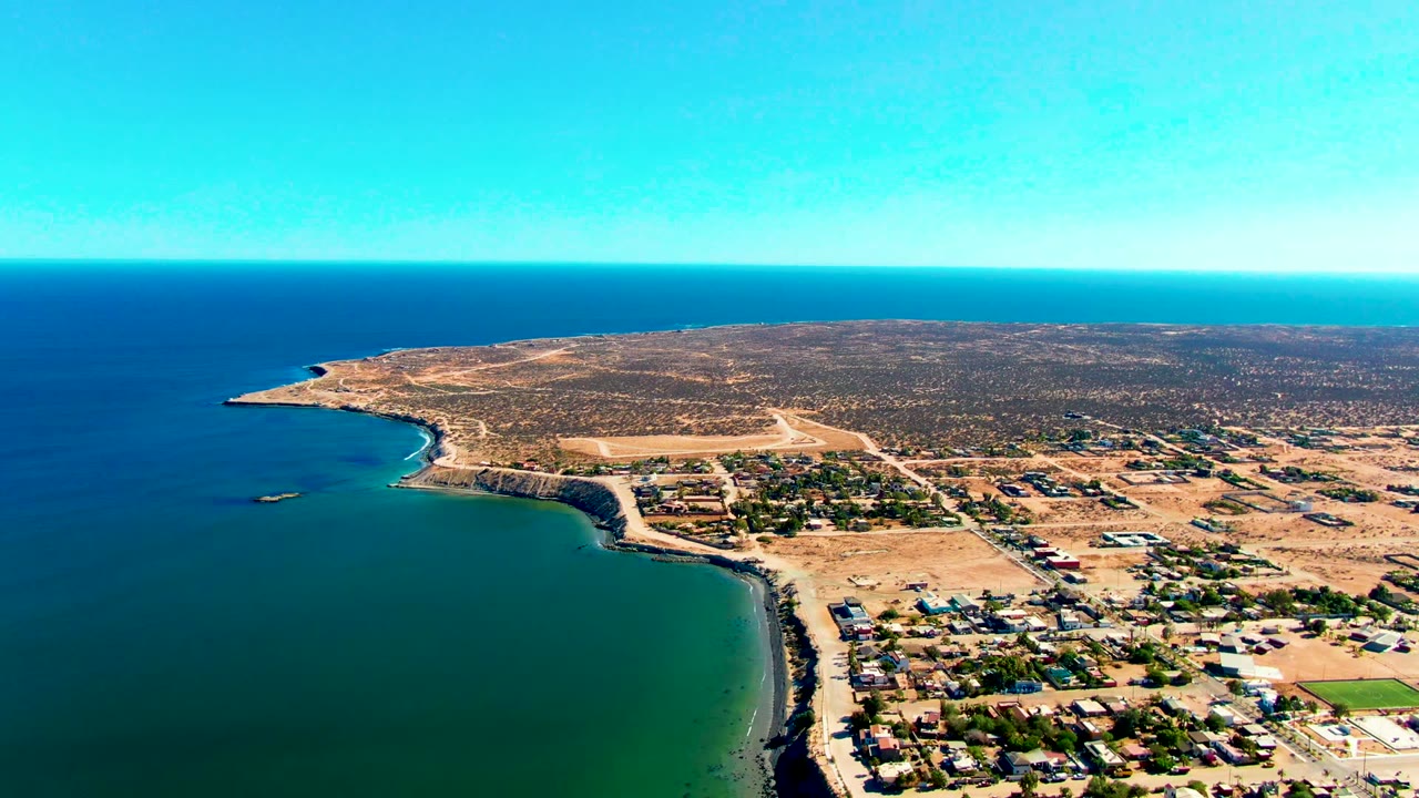 Aerial view over San Juanico, Baja California Sur, with blue ocean at left, a curved shoreline, low buildings on dry land, a small offshore boat shape, and no visible people.
