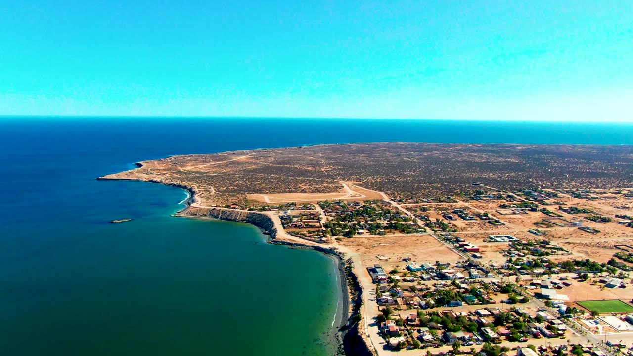 Drone view of blue water curving around a dry coastal settlement.