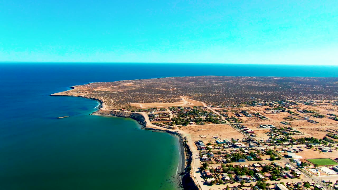 San Juanico appears from above with dark blue water beyond green shallows, a pale beach curve, scattered buildings and roads, dry inland terrain, and no visible people.