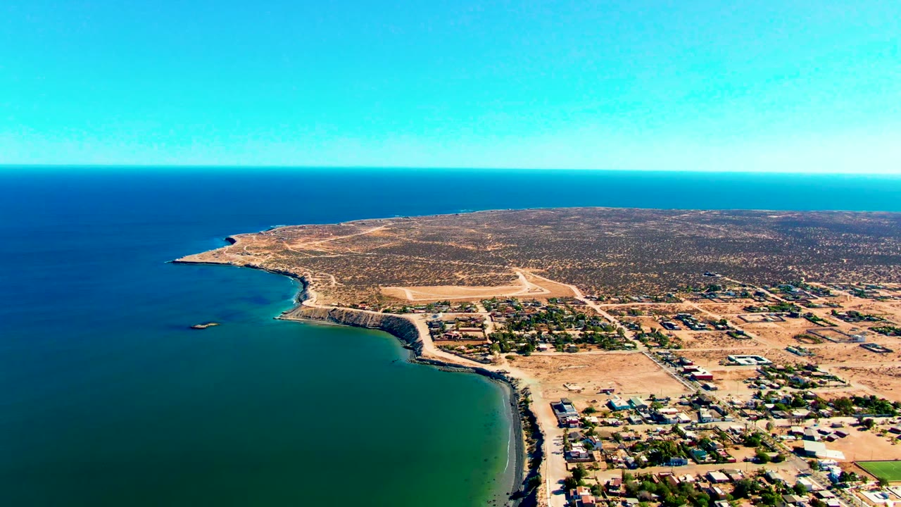 Opening aerial frame over San Juanico, Baja California Sur, showing teal nearshore water at left, a curved coast, low buildings on sandy ground, dry land beyond, and no visible people.