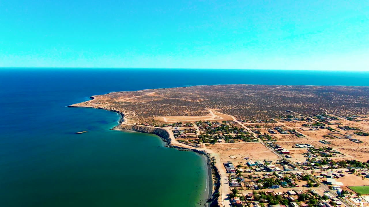 High aerial view of ocean, sandy shoreline, scattered buildings, and arid coastal terrain.