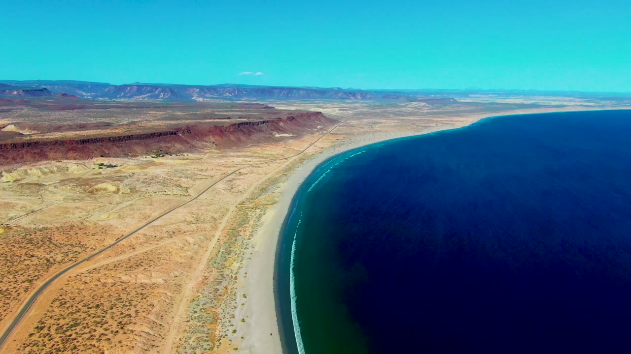 Opening aerial frame over San Juanico, Baja California Sur, showing a curving sandy beach, dark blue water, arid flats, a road, distant mesas, and no visible people.