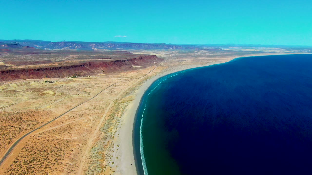 Aerial coastline view with surf along a pale beach and a road through desert.