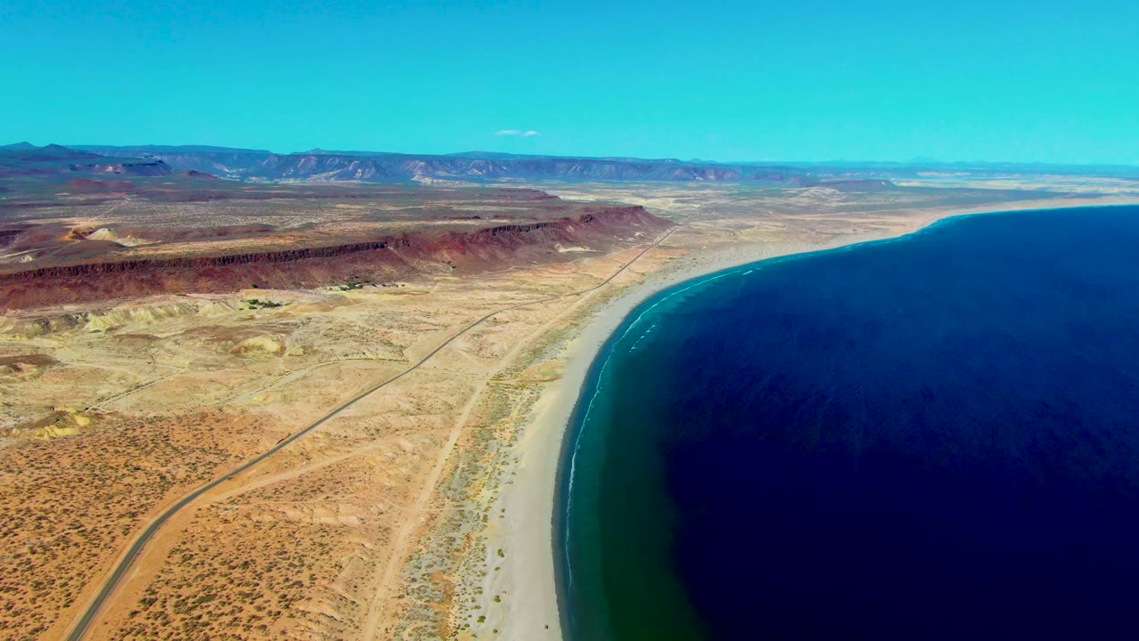 Aerial view over San Juanico, Baja California Sur, with a straight desert road, pale beach, dark blue ocean, distant mesas, and no visible people.