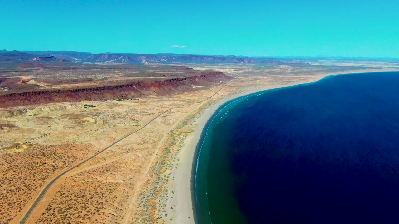 High aerial coastline with desert scrub, a road, sandy beach, and dark blue water.