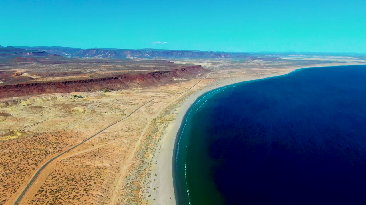 Wide aerial frame over San Juanico, Baja California Sur, with the coastline arcing through the shot, the ocean occupying the right side, and no visible people.
