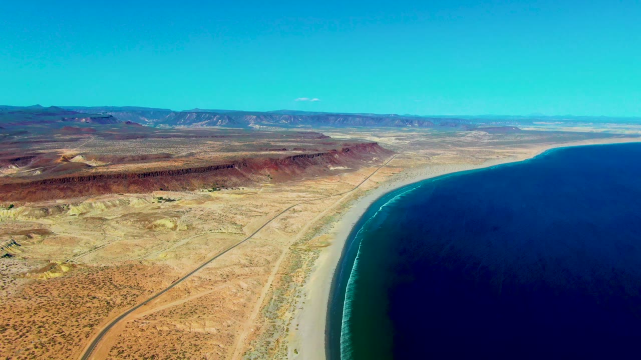 Aerial view over San Juanico, Baja California Sur, with a dirt road crossing arid ground toward a sandy beach and dark blue ocean, no visible people.