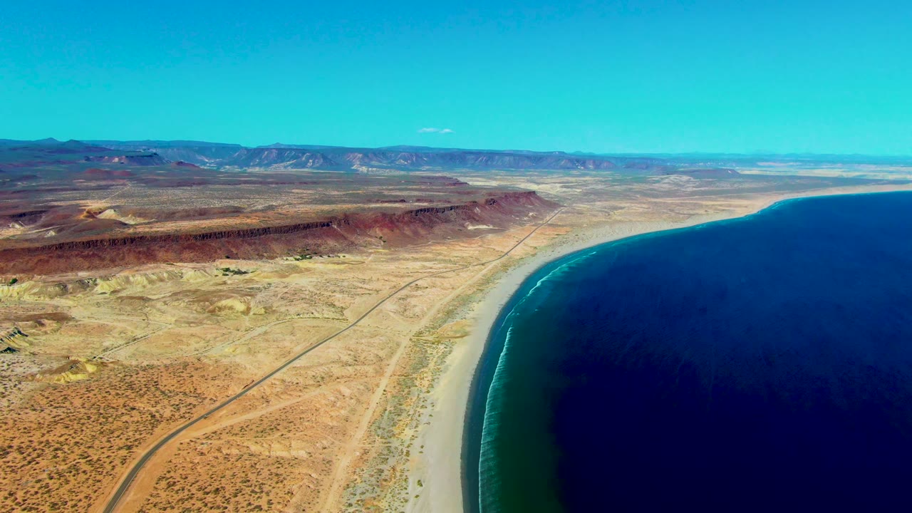 Aerial view at San Juanico with the beach bending along the right side of frame, a dirt road through dry terrain, low mountains beyond, and no visible people.