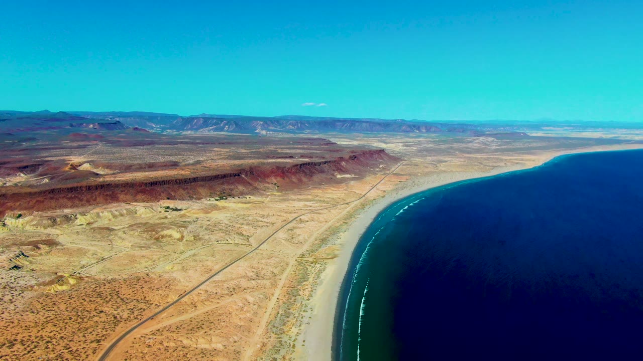 Early frame of San Juanico, Baja California Sur coastline with a dirt road through dry terrain, a curved sandy beach, dark blue ocean, distant mountains, and no visible people.