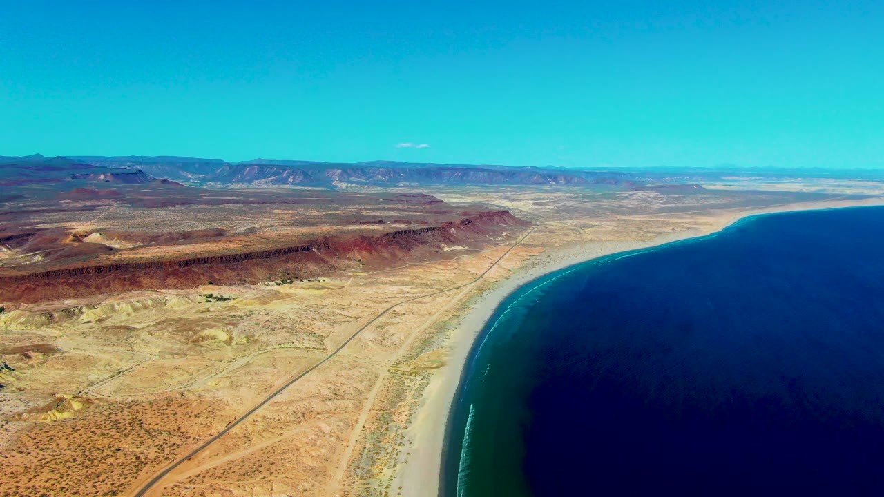 Wide aerial coastline with road, sand, ocean, and mountains.