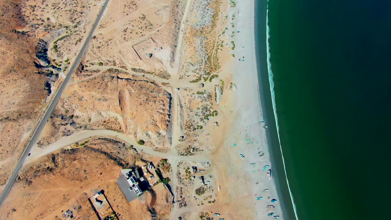 Overhead view of San Juanico beach with pale sand, scrubby dunes, a diagonal road, small beach umbrellas, tiny visible people, and green ocean on the right.