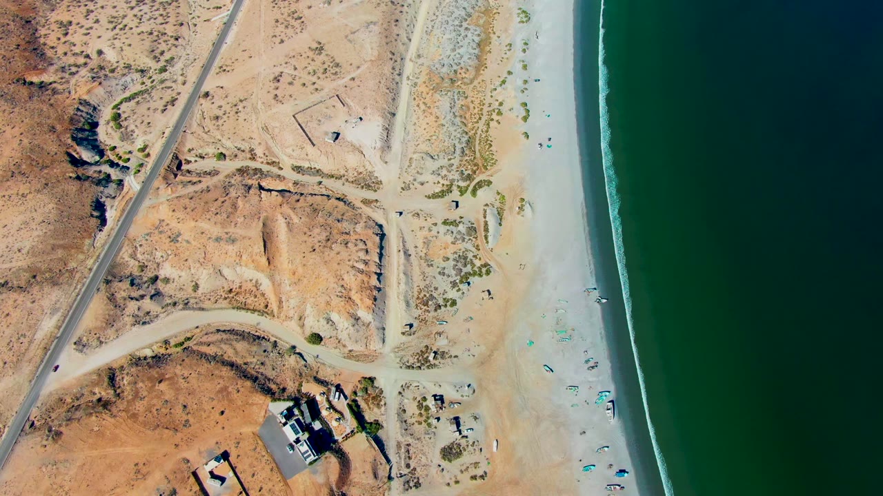 Aerial frame over San Juanico showing the road and dry dunes to the left, scattered umbrellas and tiny visible people on the sand, and a dark green ocean band to the right.