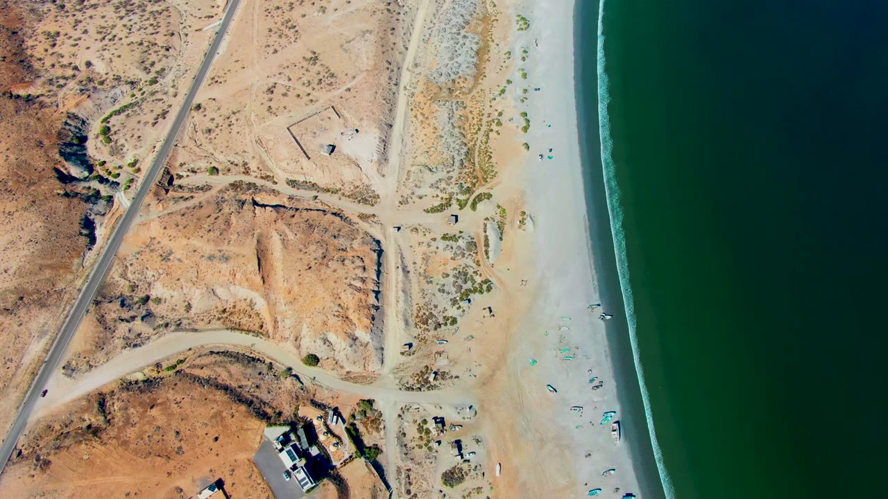 Top-down San Juanico coastal view with a diagonal paved road, pale dune patches, small beach setups, tiny visible people, and green water bordered by a narrow surf strip.