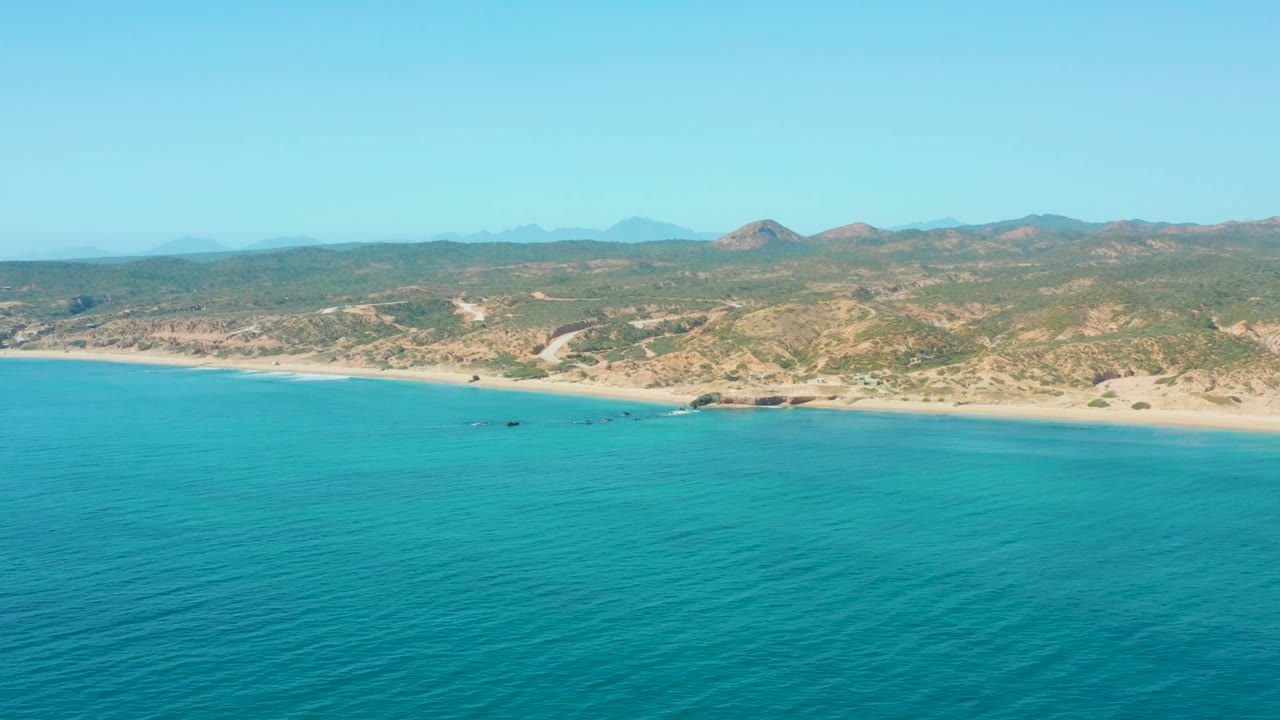 Aerial view of El Tule Los Cabos with turquoise ocean in the foreground, sandy shoreline and low rocks near the beach, distant hills under clear sky, and no visible people.