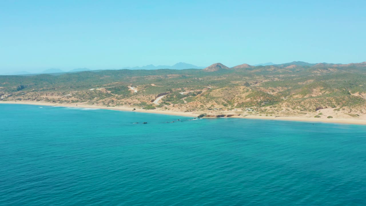 El Tule Los Cabos coastline seen from above with turquoise water across the lower frame, sand and rocks along shore, arid hills beyond, a clear horizon, and no visible people.