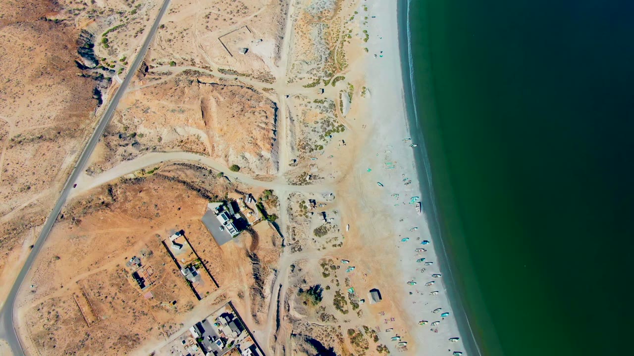 Opening aerial frame over San Juanico, Baja California Sur, with a road crossing dry desert terrain, small buildings near the sand, beach umbrellas along the shoreline, and no visible people.