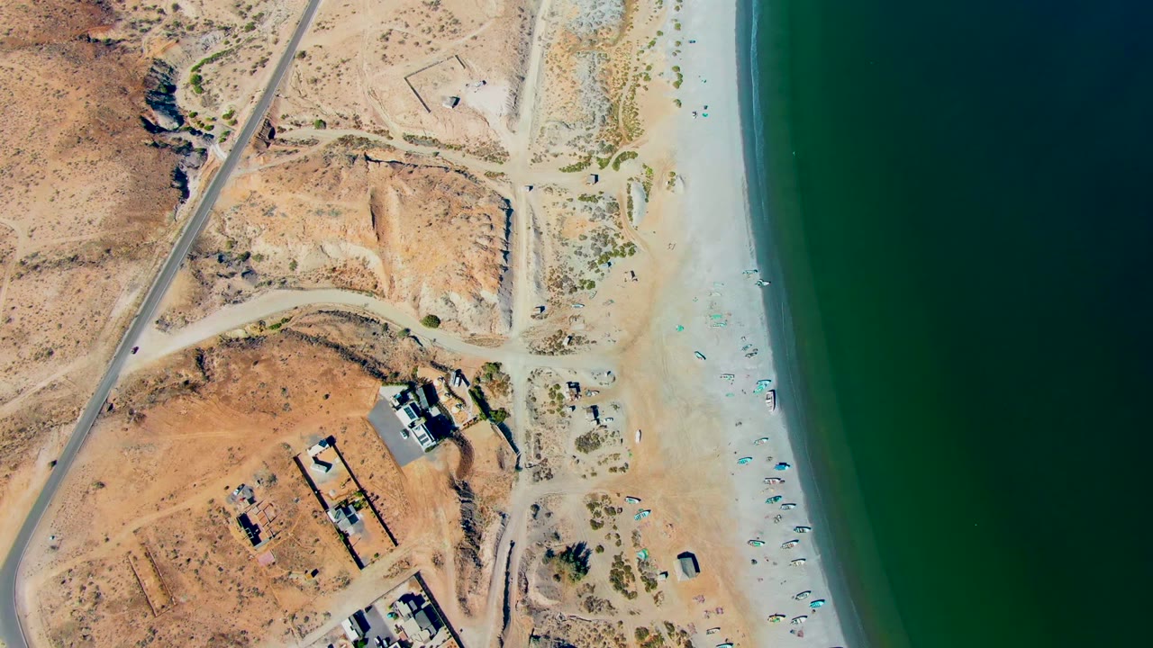 Overhead coastal road beside desert lots, beach umbrellas, shoreline, and ocean.