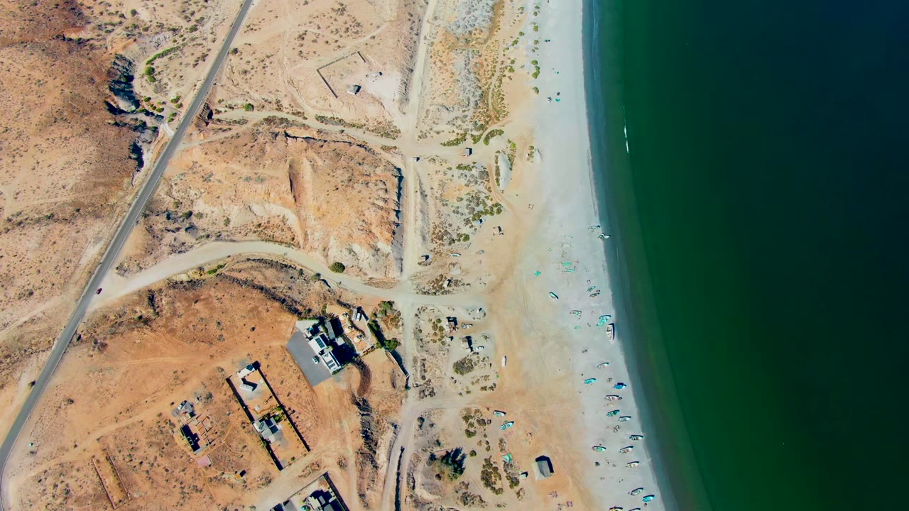 San Juanico aerial frame with the road running through arid ground, beach umbrellas dotting the sand, a narrow surf line at the water edge, and no visible people.