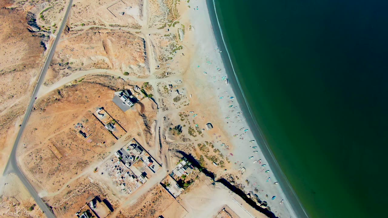 Opening overhead frame in San Juanico, Baja California Sur showing arid ground and road at left, scattered construction structures near the beach, green ocean at right, and no visible people.