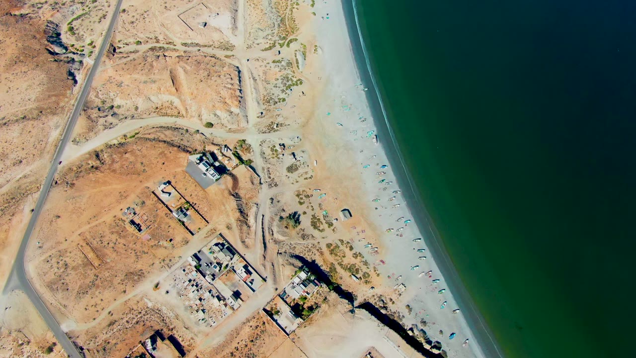 Aerial shoreline view with road, sandy beach, buildings, and dark green ocean.