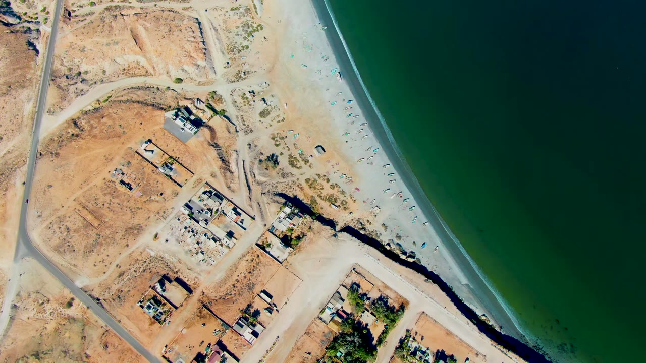Overhead view in San Juanico, Baja California Sur showing dry lots, a coastal road, scattered buildings, pale beach, green water, and no visible people.