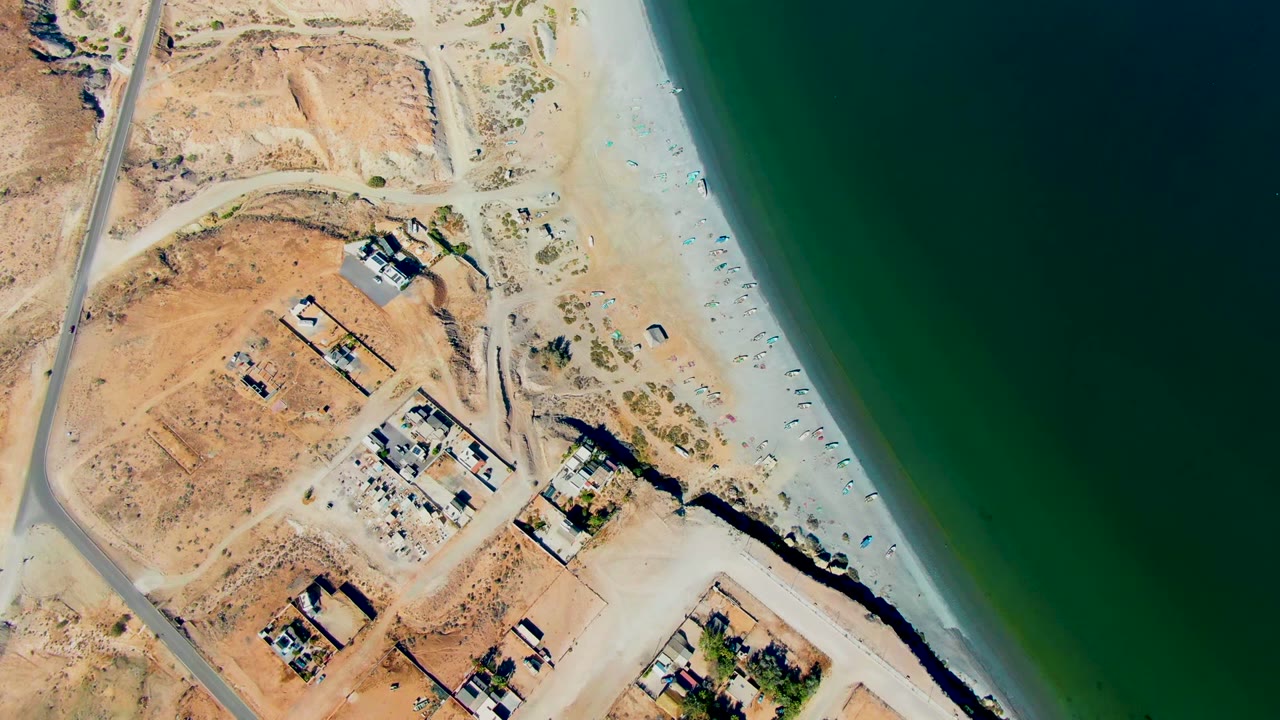 Overhead view in San Juanico, Baja California Sur, showing dry terrain, a coastal road, scattered buildings, sandy beach, dark green water, and no visible people at the start of the clip.