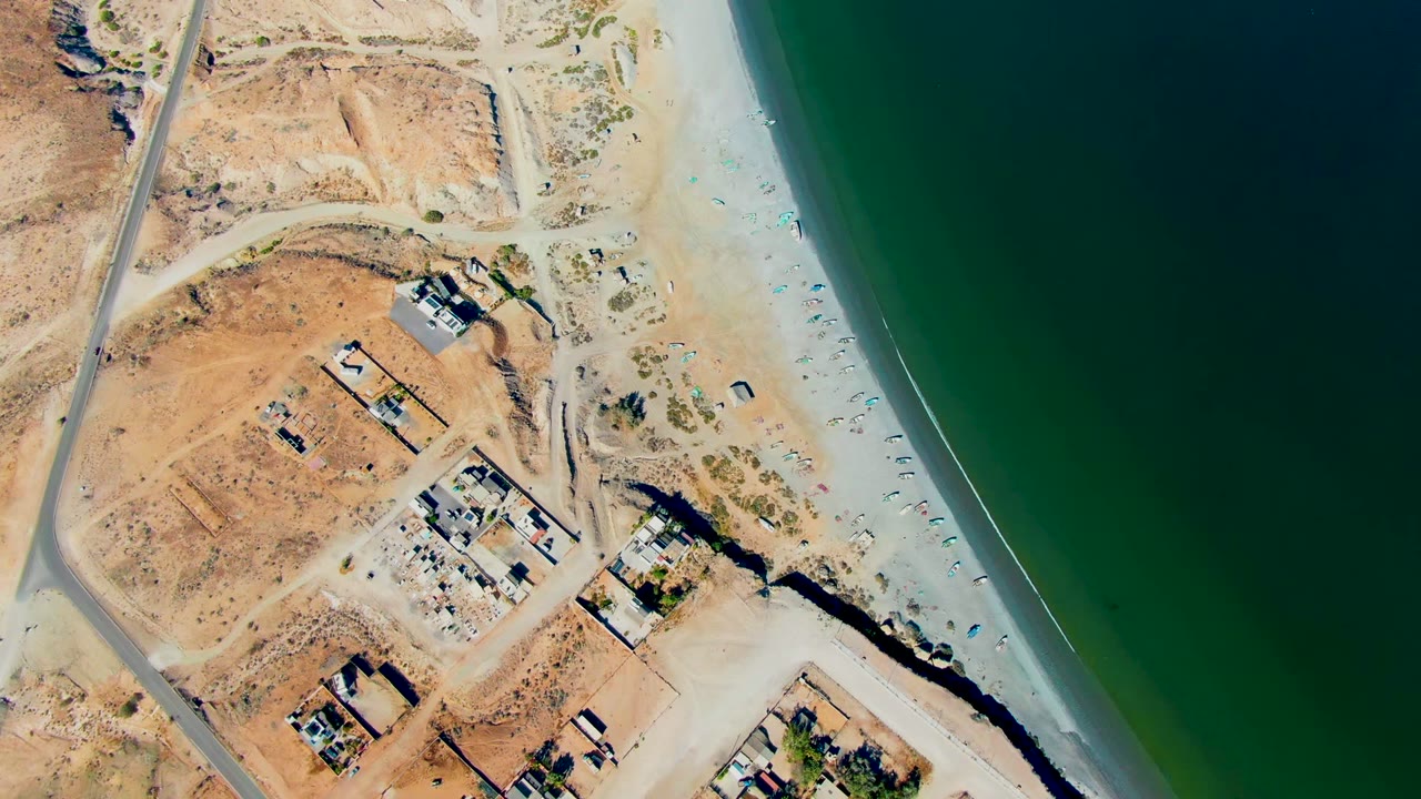 Top-down view in San Juanico, Baja California Sur, showing tan dry terrain and small structures on the left, a narrow sandy beach, dark green ocean water on the right, and no visible people.