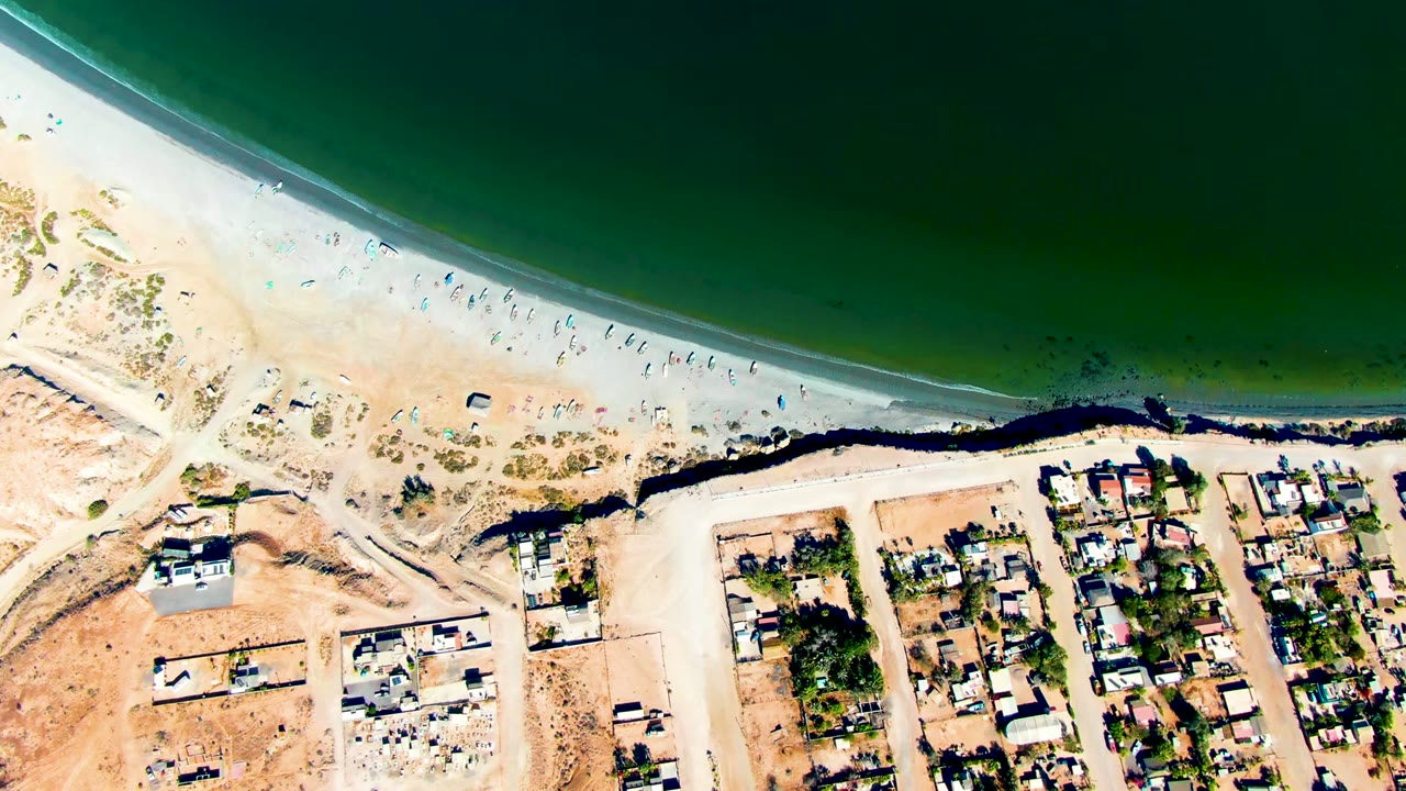 Aerial view of San Juanico, Baja California Sur with green ocean water, pale sand, scattered beachgoers, a dark shoreline edge, roads, and residential buildings.