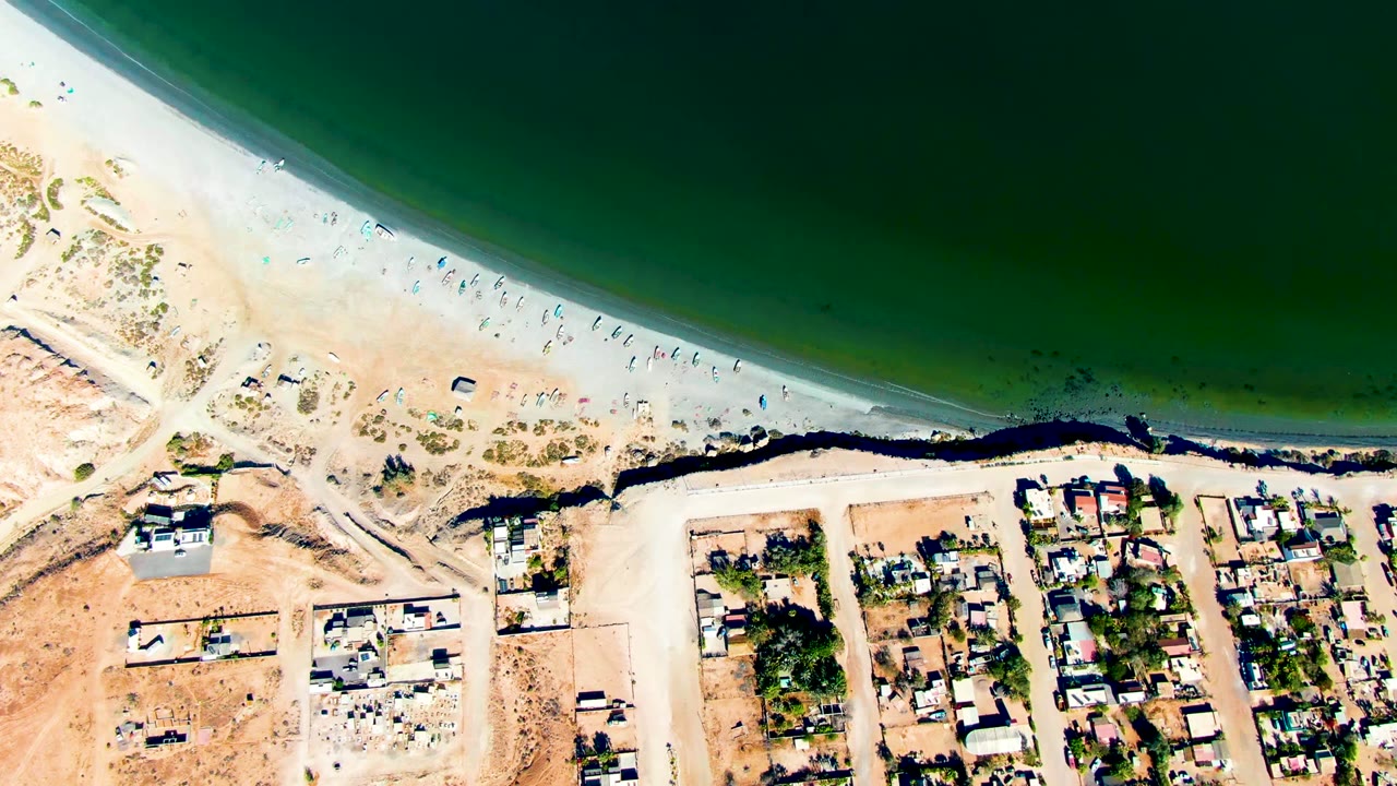 Overhead view in San Juanico, Baja California Sur with green water across the top, a pale beach with visible people, and compact residential blocks below the shore.