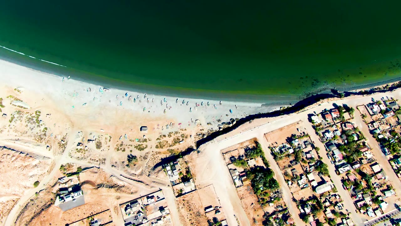 Aerial frame of San Juanico where the pale beach curves below green water, with scattered beachgoers, a dark shoreline band, and homes at the right.