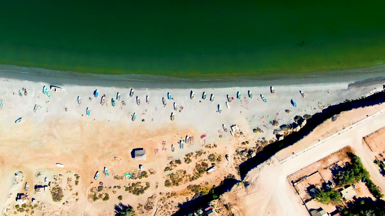 Overhead view of San Juanico, Baja California Sur, with green water along the top edge, small boats on pale sand, scattered umbrellas and vehicles, and a dark channel cutting through the beach.
