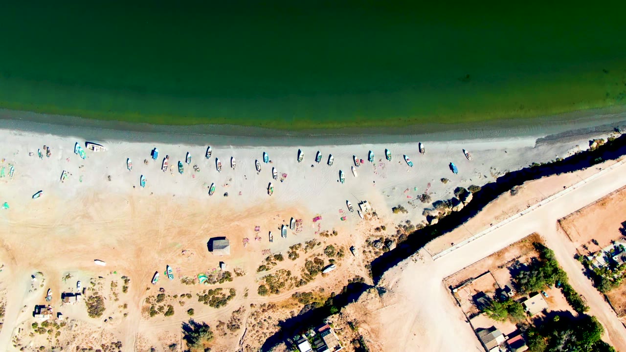 Aerial shoreline with boats lined along the sand below green water.