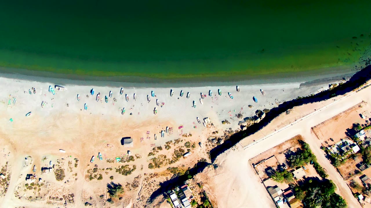 Top-down view in San Juanico with green water, pale beach, small boats scattered along the shore, a dark channel or seawall, and nearby buildings at the lower right.