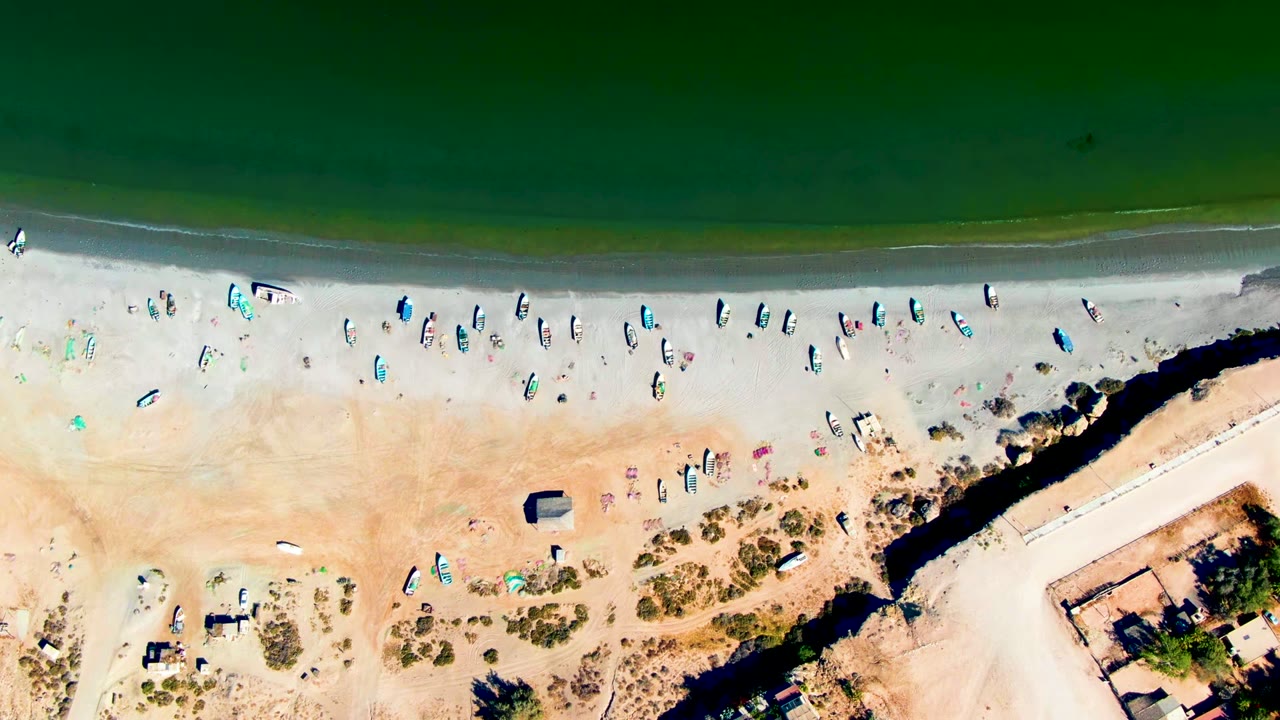 Overhead view of San Juanico beach in Baja California Sur, Mexico, with green water at the top, a pale sandy shoreline, scattered beachgoers, small boats or boards, and scrubby sand below.