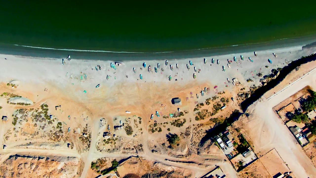 Top-down aerial frame over San Juanico, Baja California Sur, with green ocean at the top, small boats on the sand, scattered beachgoers, a road, and low buildings near the lower right.