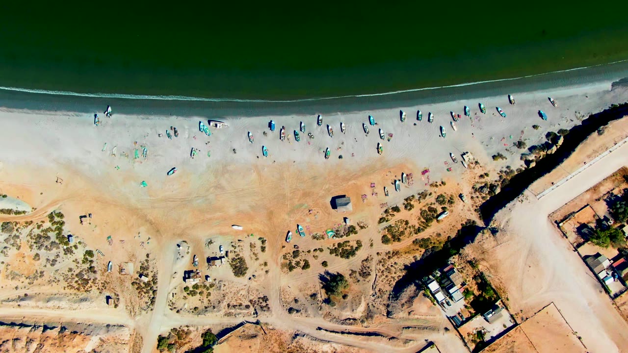 Overhead beach scene with boats lined near the water and a sandy road at right.