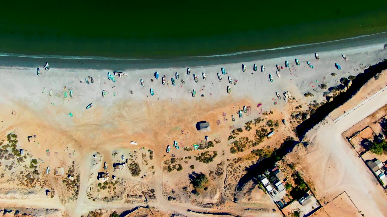 San Juanico aerial frame with green water across the upper edge, a bright sandy strip dotted with boats and beachgoers, and tan access roads and structures at the right side.