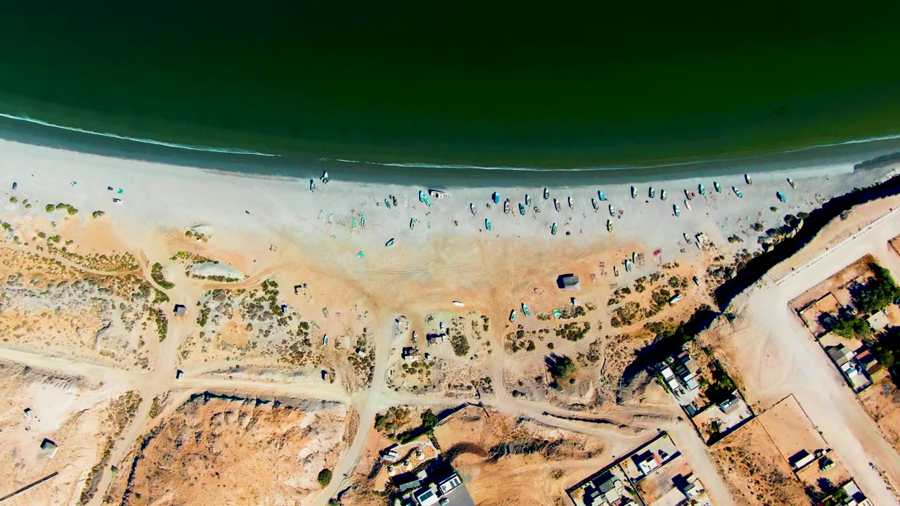 Straight-down aerial frame over San Juanico, Baja California Sur, with dark green ocean above a pale sandy beach dotted with tiny people and gear near dunes and buildings.