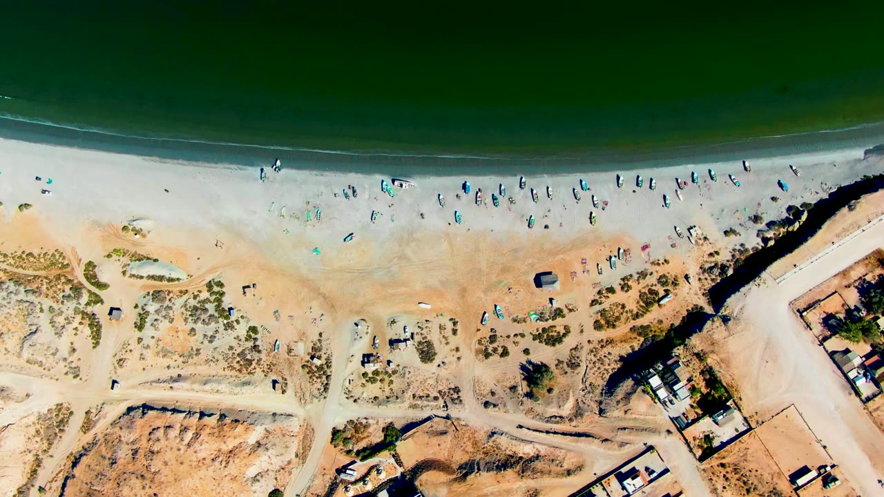 San Juanico overhead frame centered near a dark breakwater or jetty, with beachgoers spread along the sand and roads and buildings bordering the dune area.