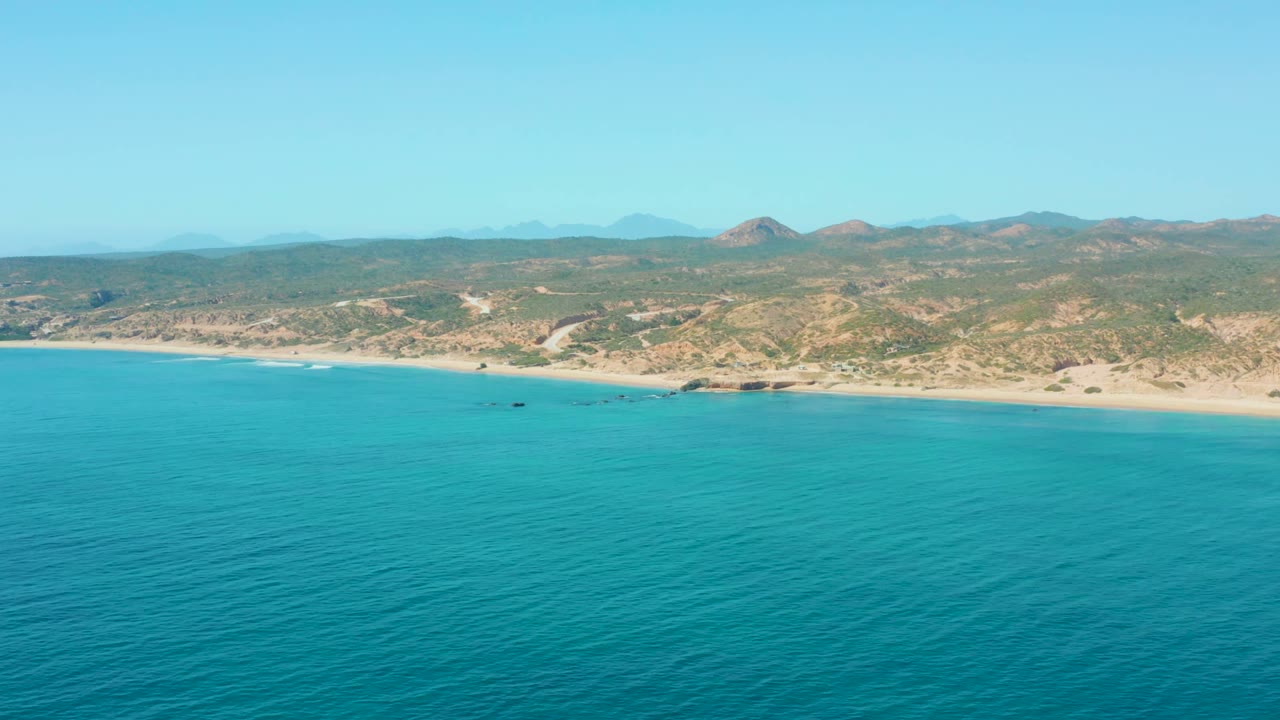 Aerial frame over El Tule Los Cabos showing turquoise water in the foreground, a pale sandy beach, scrubby hills, small boats near shore, clear sky, and no visible people.