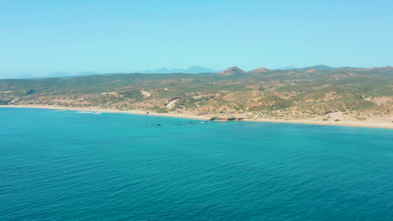 Wide aerial coastline with calm blue water, sandy beach, hills, and small boats offshore.