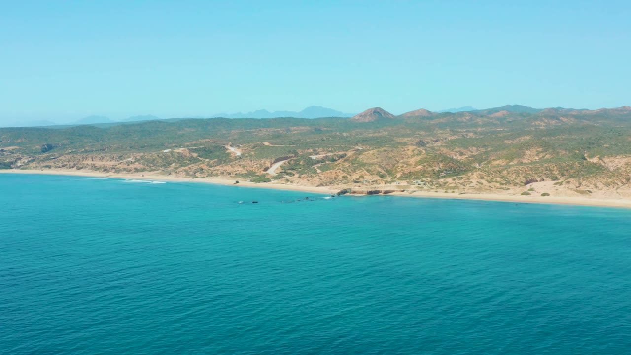 El Tule Los Cabos coastline appears from a high offshore view, with textured turquoise water, pale sand, sparse vegetation on the hills, distant mountains, and no visible people.