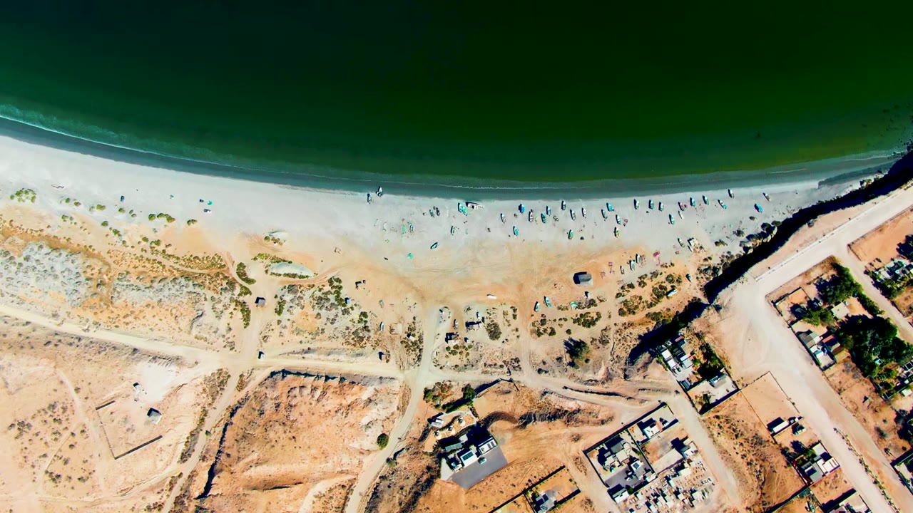 Overhead view of San Juanico beach with green water at the top, a pale sandy shore, scattered beachgoers near the surf, and roads and buildings below.