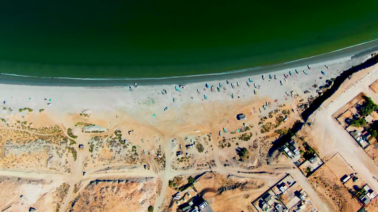 Aerial view over San Juanico, Baja California Sur showing green ocean, pale shoreline, scattered beachgoers, umbrellas, dunes, parked vehicles, and inland structures at the start of the clip.
