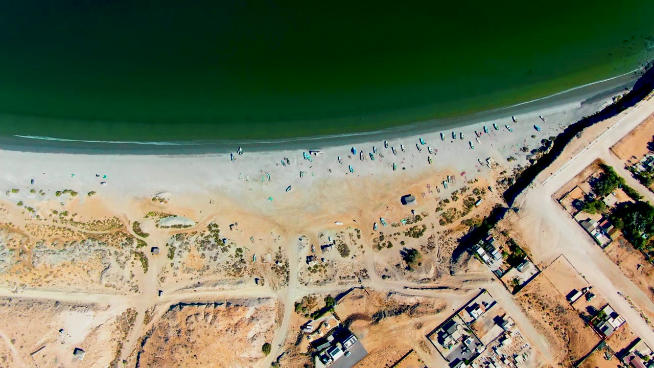 High aerial shoreline view with scattered beachgoers, umbrellas, dunes, vehicles, and buildings.