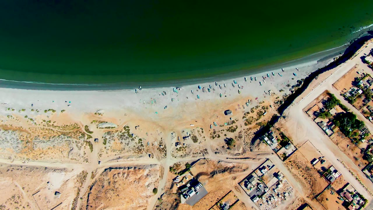 San Juanico appears from above with the dark green ocean at the top, a thin white surf line, beachgoers and umbrellas along the sand, and a diagonal dirt road beside nearby structures.
