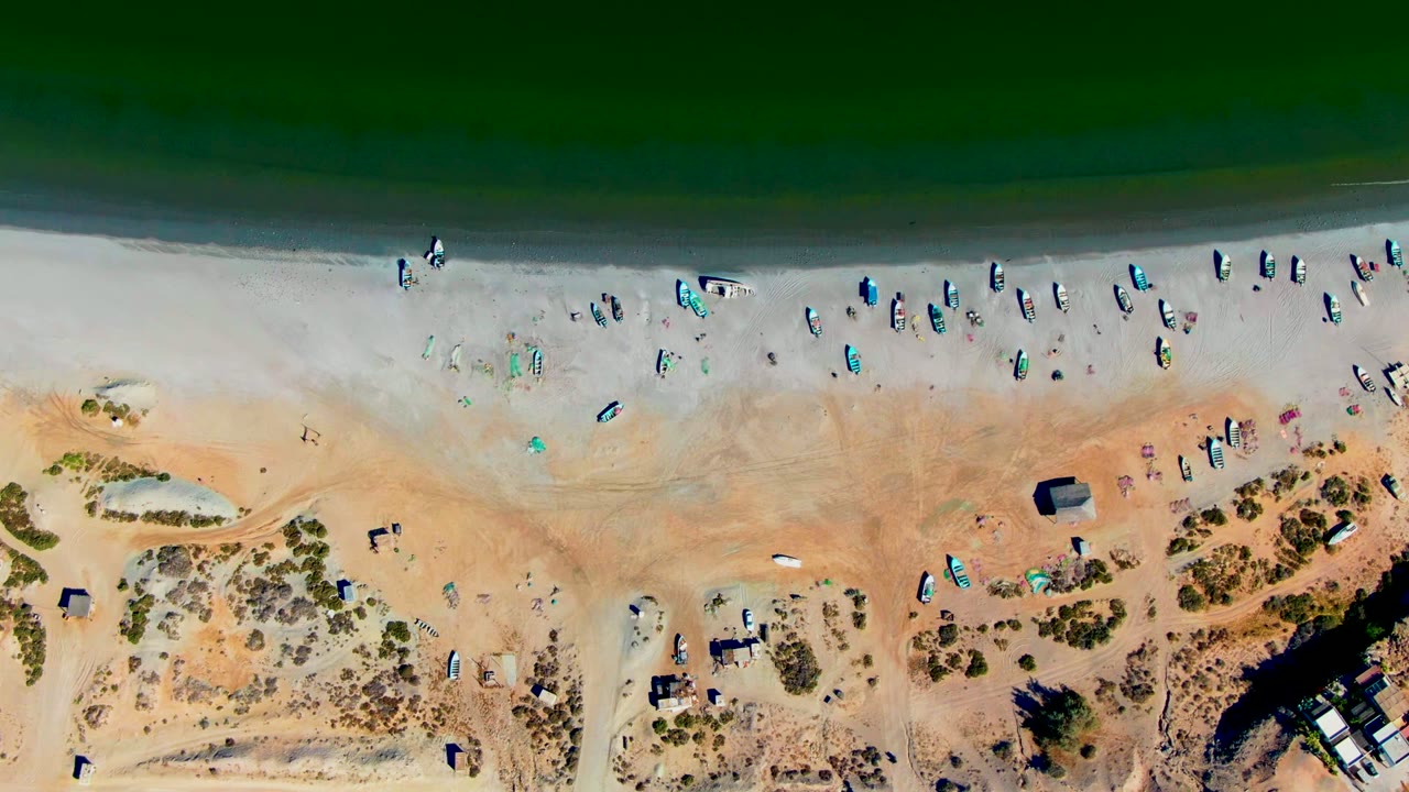 Top-down San Juanico beach view with a dark water band, narrow surf edge, scattered people and gear on the sand, dune shrubs, paths, and a few small structures near the lower frame.