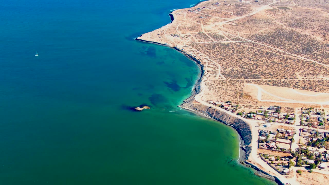 Aerial frame over San Juanico, Baja California Sur, showing a small sailboat at left, green shallows near a rocky point, desert roads, shoreline buildings, and no visible people.